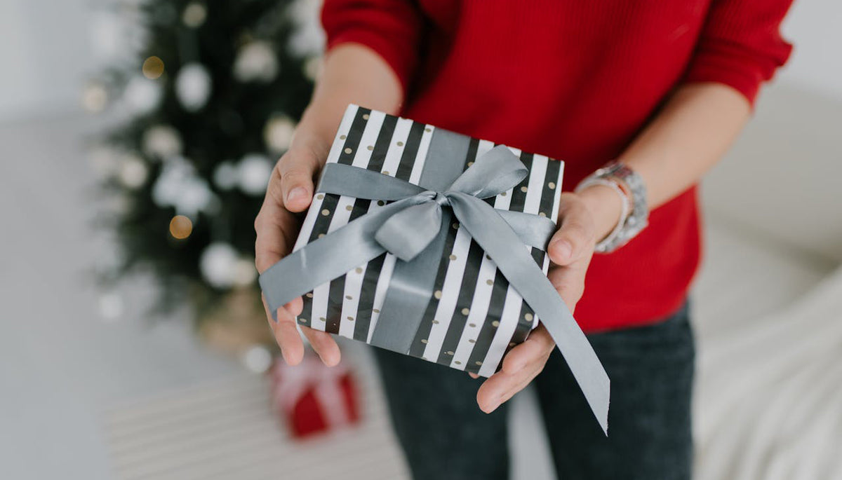 close up shot of a woman wearing a red sweater holding a gift wrapped in striped wrapper there is a small Christmas tree in the background