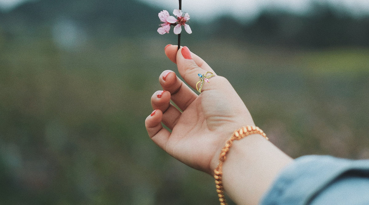 woman's hand wearing a gold-plated stainless steel ring with pastel pink and blue cz stones holding up a pink flower