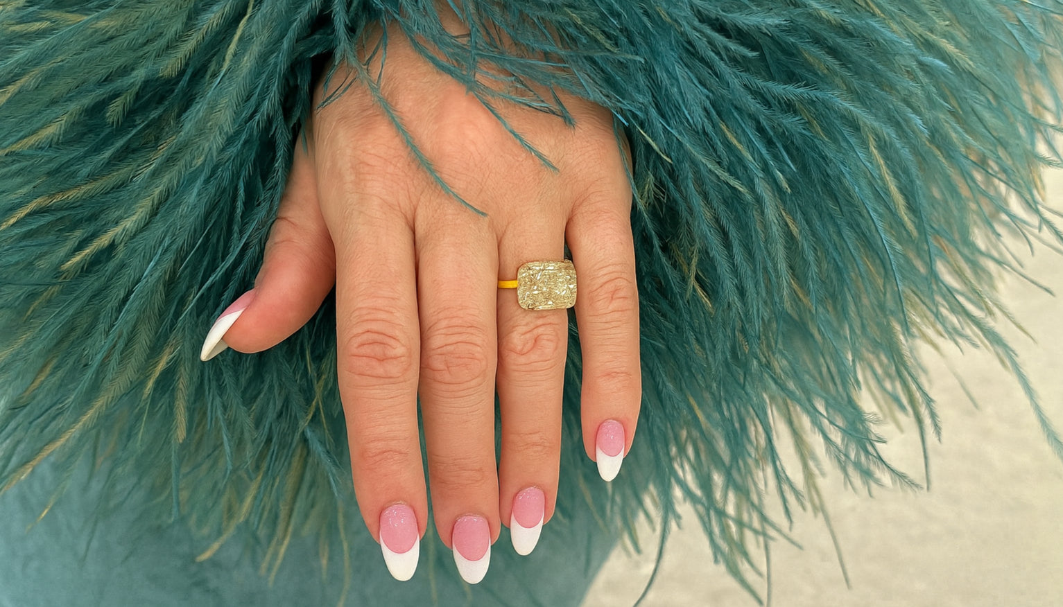 close up shot of a hand with long french tip almond nails wearing an east-west engagement ring and a dress with teal feather embellisments