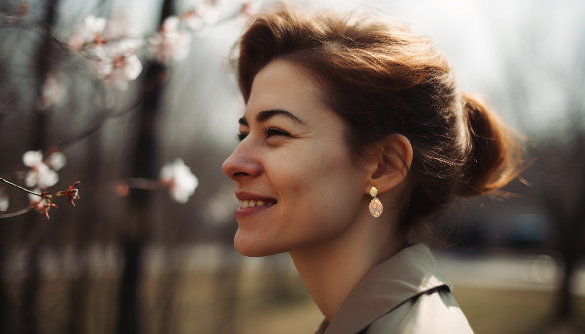 side profile a smiling brunette with her hair in a bun wearing CeriJewelry's Gold-Plated Stainless Steel Pastel Pink Epoxy Drop Earrings with cherry blossoms in the background